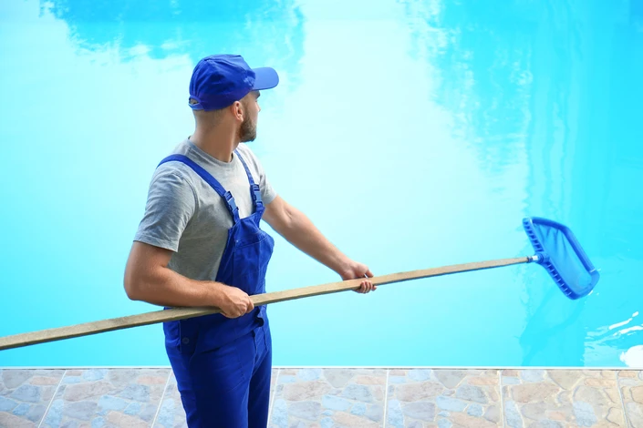 Un technicien en combinaison bleue nettoie une piscine extérieure avec une épuisette à manche télescopique