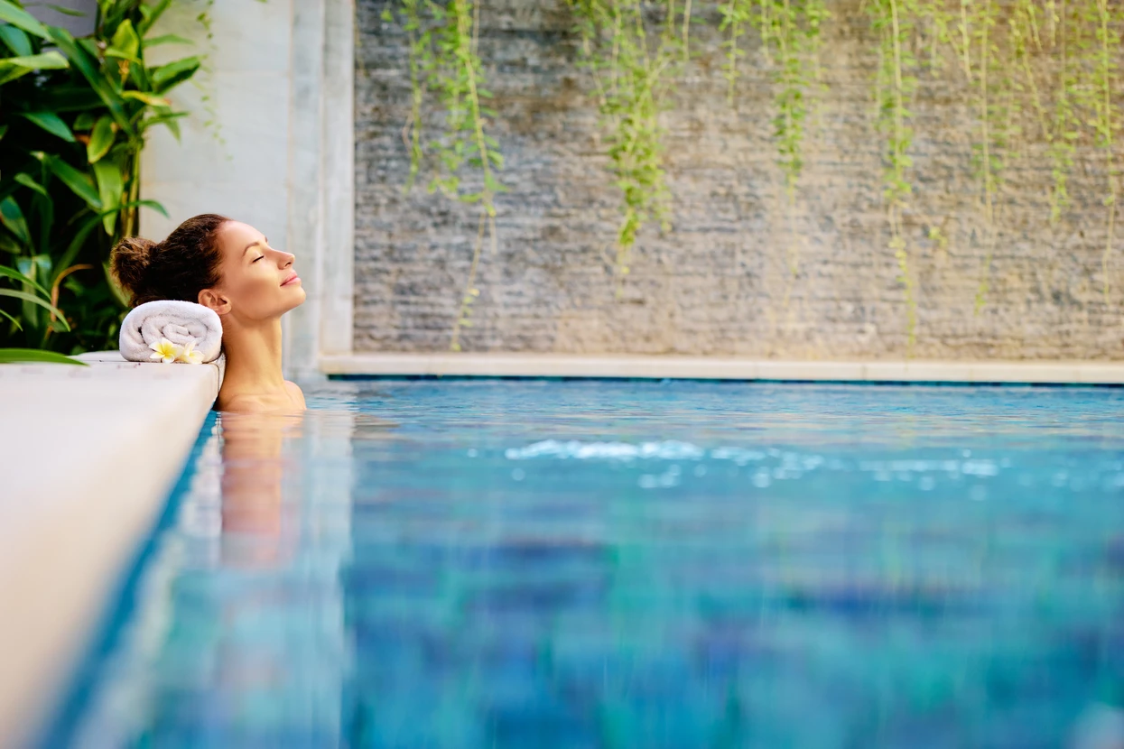 Femme en maillot de bain et chapeau blanc se détendant au bord d'une piscine, jambes dans l'eau cristalline